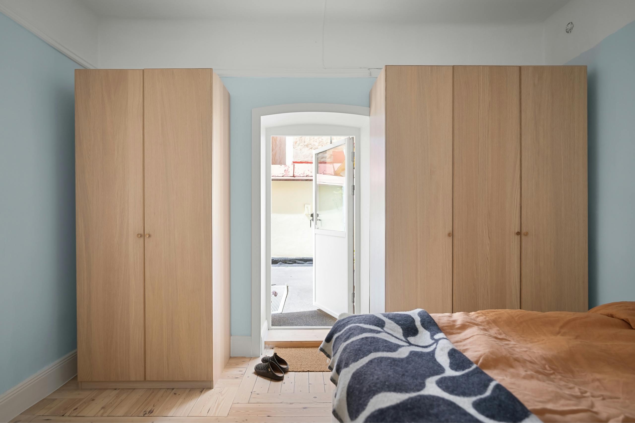 A minimalist bedroom with sunlight streaming through a door, featuring wooden wardrobes and a cozy bed with colorful blankets.